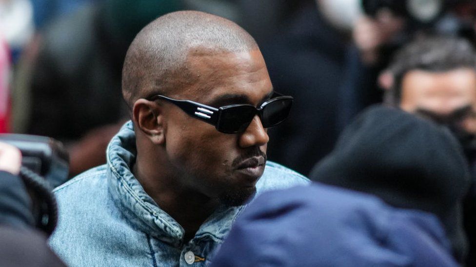 Kanye West, a black man with a shaved head, is pictured at Paris Fashion Week in January 2023. He is wearing dark sunglasses and a denim effect puffer jacket. He is not looking at the camera and looks to the right - he has a short moustache and goatee.