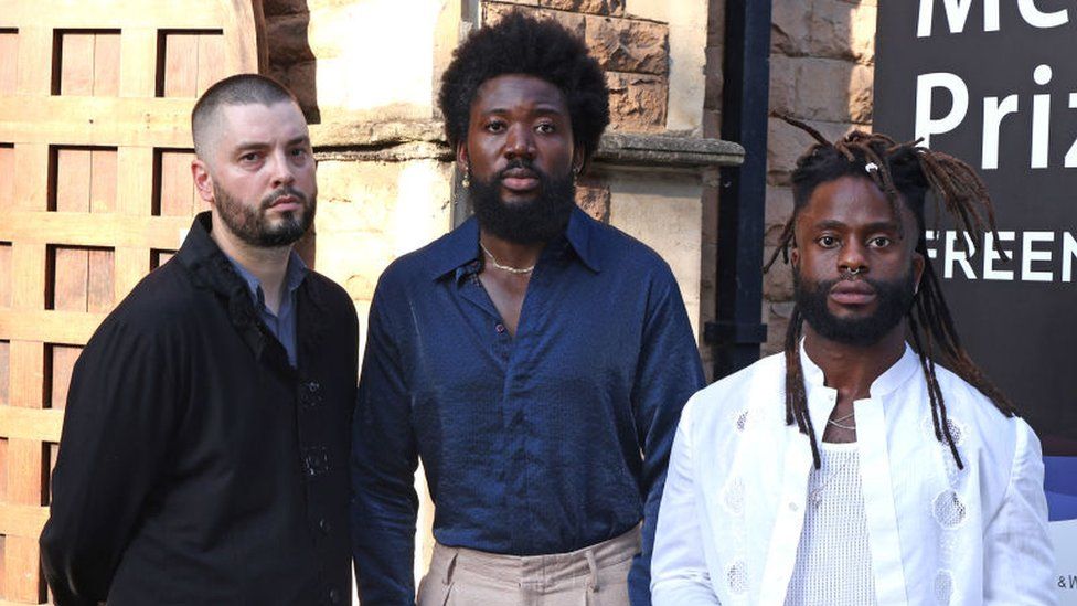 Graham Hastings, Alloysious Massaquoi and Kayus Bankole of Young Fathers attends The Mercury Prize 2023 awards show at Eventim Apollo on September 07, 2023 in London. Graham is a white man in his 30s with shaved dark hair and a short dark beard. He wears a black jacket over a blue shirt. Alloysious stands beside him - he is a slightly taller black man also in his 30s. He has afro hair and a beard about two inches long. He wears a blue shirt with the top two buttons undone, revealing his chest and a gold chain necklace. Beside him is Kayus, who is the shortest of the three artists. He is a black man with his hair styled in dreads and a short beard. He wears a white shirt open over a white vest with three necklace chains and a nose ring