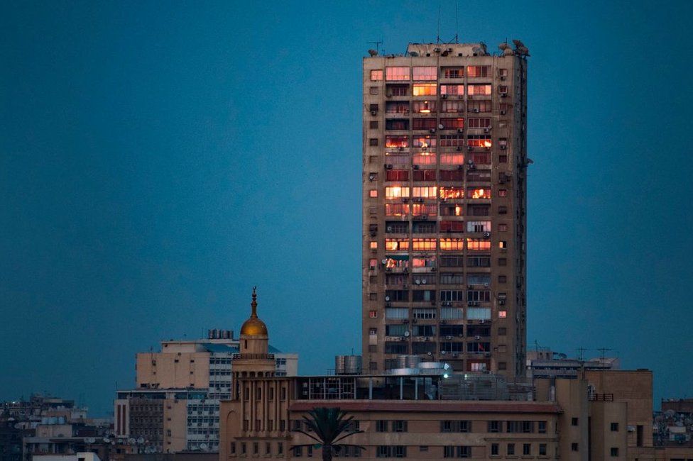 A tall tower block stands next to a domed mosque in a built-up part of the city of Cairo. The sky is a dark blue and the tower's windows are illuminated by the sun in a bright orange. Egypt - Wednesday 20 February 2019