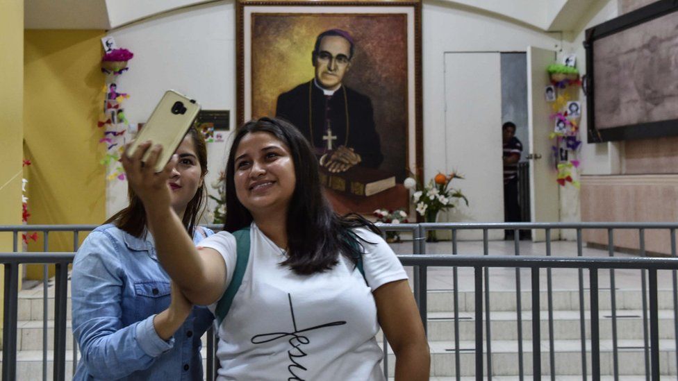 Two women take a selfie with a portrait of Archbishop Oscar Romero