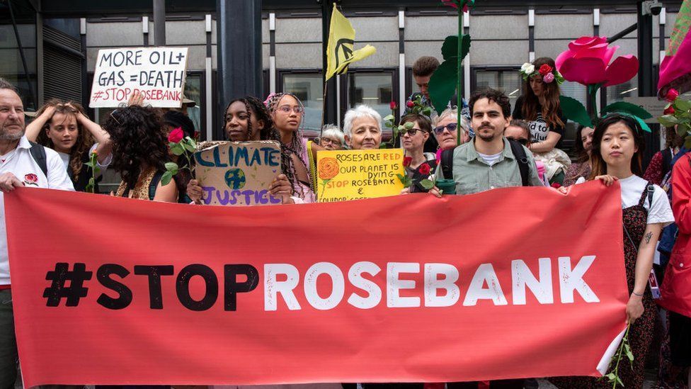 Group of protestors holding small signs standing behind a large red stop rosebank banner.