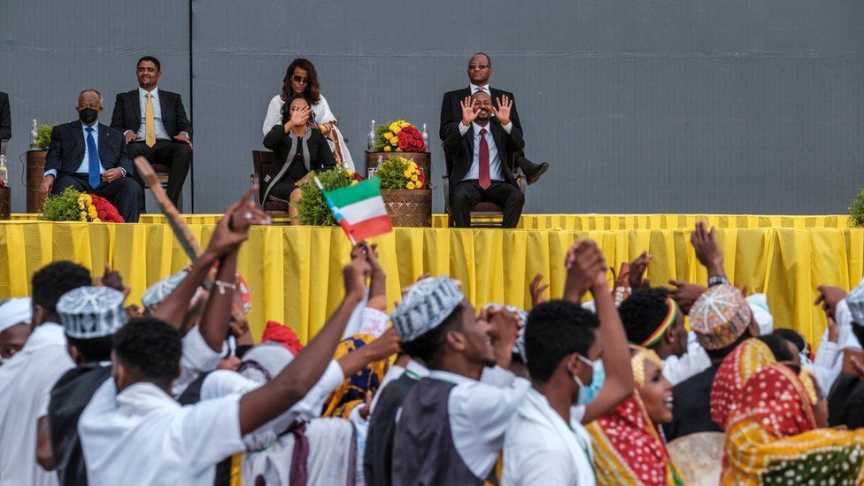 Ethiopia's newly sworn in Prime Minister waving to a crowd of people dressed in traditional clothing. One person is waving an Ethiopia flag. Abiy Ahmed is on an elevated stage with yellow draping. He is also accompanied with other people on the stage.