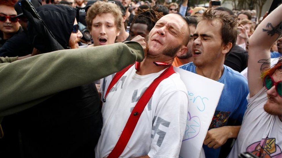 A man wearing a shirt with swastikas is punched by a member of a crowd of protesters at University of Florida's campus, where white nationalist Richard Spencer gave a speech.