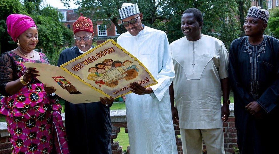 A photograph tweeted by the @NGRPresident Twitter account shows President Buhari standing outdoors, grinning and wearing sunglasses while holding a large get well card, with a handful of officials around him.