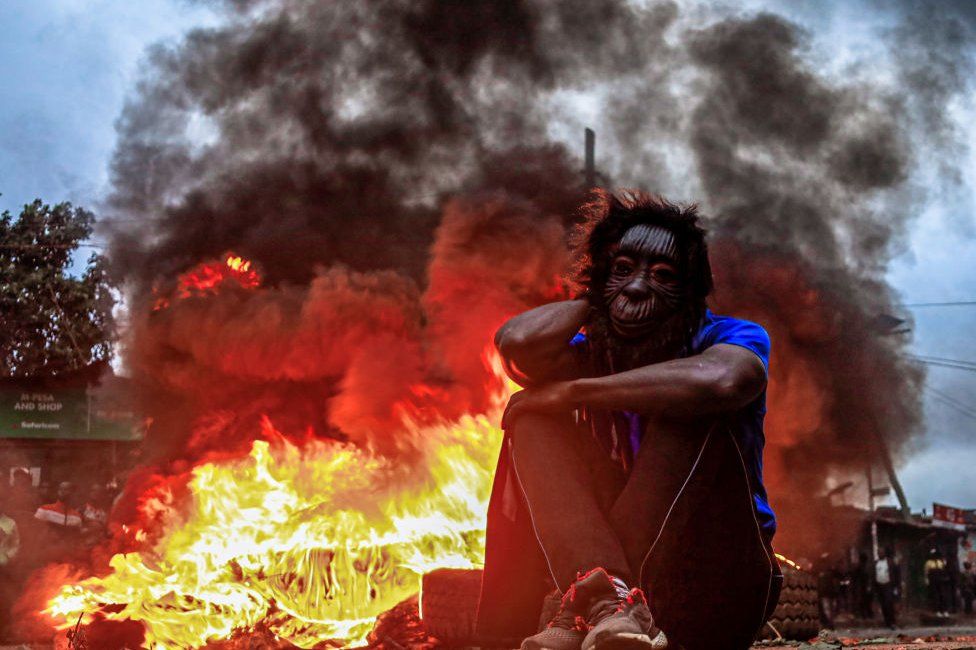 A man with a cat-like mask sitting on the ground in front of a huge blaze.