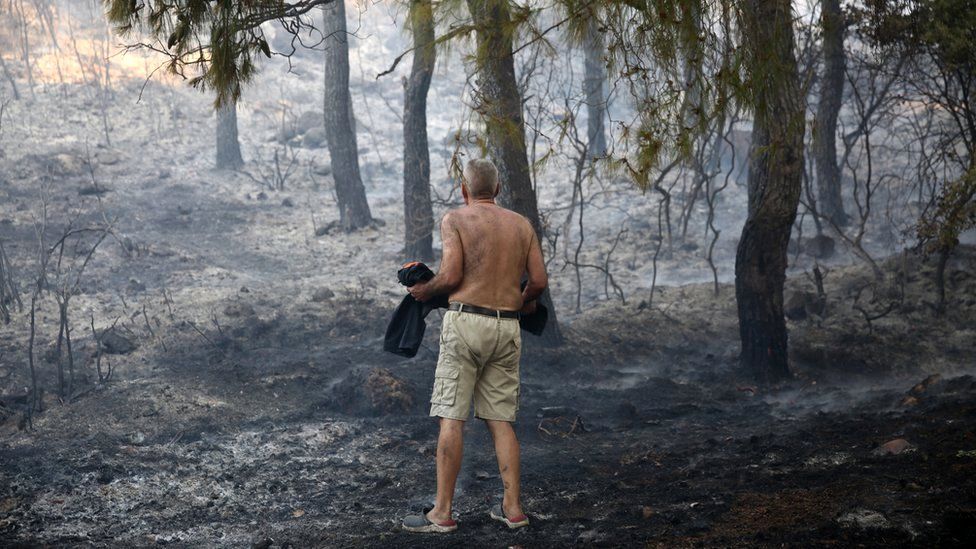 A resident looks at the burned area as he carries a burning tree trunk during a wildfire in Thrakomakedones area, near Athens, Greece, 7 August 2021