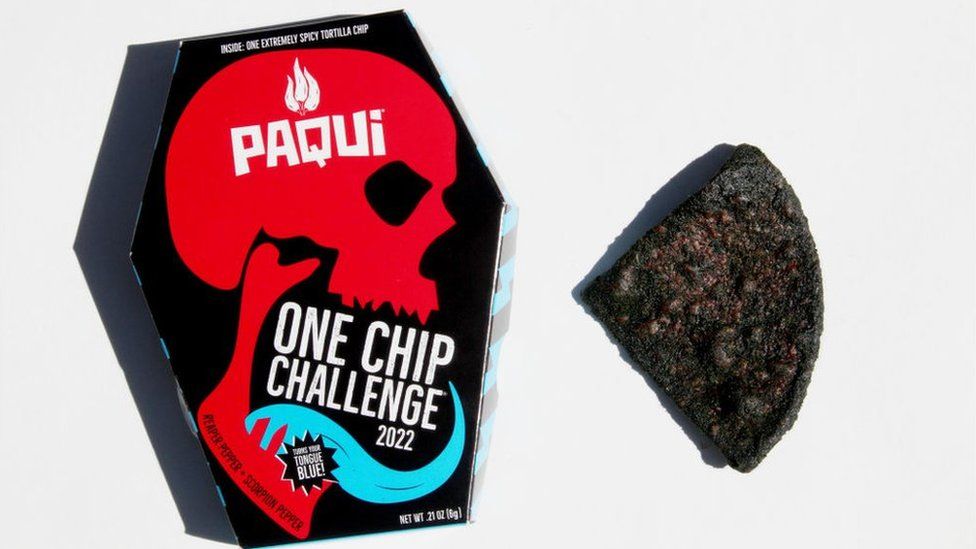 A coffin-shaped box and a large, black tortilla chip on a plain white background. The box is printed with a red skull in profile, with a long blue tongue emerging from its mouth. At the top of the box it says "Inside: One extremely spicy tortilla chip". Inside the skull's forehead, in white, is the company name Paqui, in white font, with three inverted peppers above it, representing a fire shape. "One Chip Challenge" and "Turns your tongue blue" also appear on the box, as do the words "reaper pepper" and "scorpion pepper".