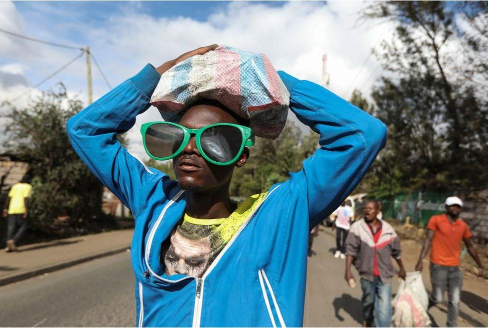 Man wearing giant green sunglasses carrying a sack of stones on his head as he stands outside.