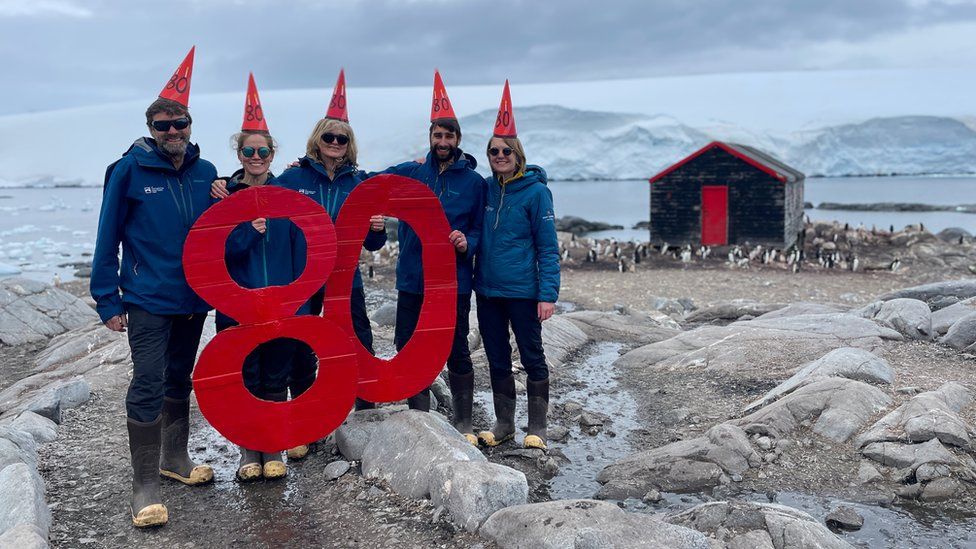 Five people in blue coats and sunglasses standing in Antarctica holding a giant red '80' sign and wearing red pointed hats. There is a shack with a red door in the background and faint mountains beyond that. There is no snow on the ground, just rocks.