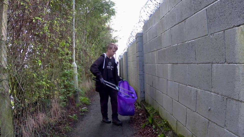 A young man in an alley. He is wearing black clothing, holding a large purple plastic litter bag and a pair of long tongs to put objects in the bag