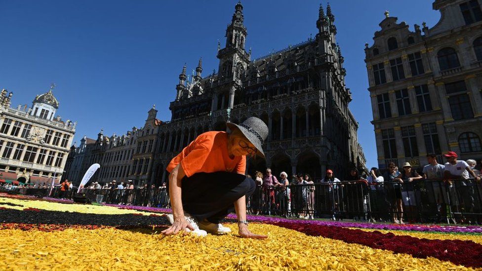 A woman wearing a hat and glasses carefully places flowers in the carpet as people watch