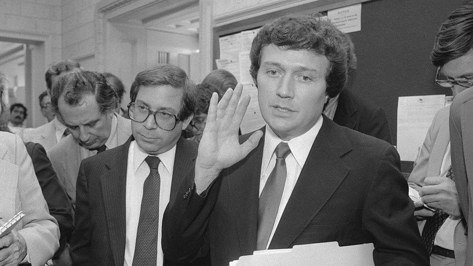 Black and white photo of man in dark suit raising his hand, surrounded by other men.