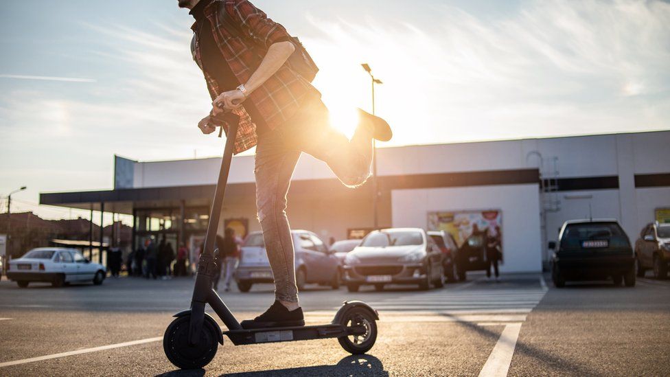 A stock photo of a man using an e-scooter in a car-park with the sun flaring behind him