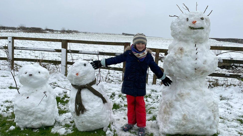 A child stands in the middle of a row of three snowmen with their arms held out to the side