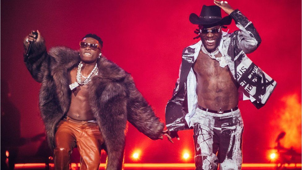 Wizkid and Burna boy performing on stage. Burna Boy is smiling and wearing a cowboy hat. Wizkid is wearing a big fur coat. Both have on sunglasses and jewellery. There is a red background.
