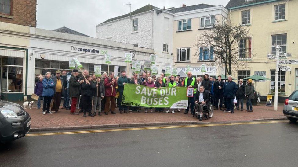 The crowd gathered in Wiveliscombe with a 'Save our Buses' banner