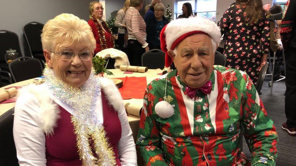 Woman wearing tinsel sits next to a man wearing a Christmas hat and sweater