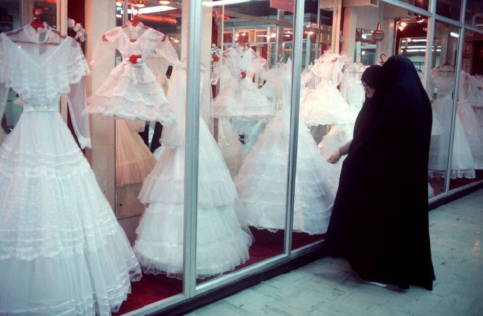 Two women wearing black chadors look at wedding dresses through glass windows in 1986