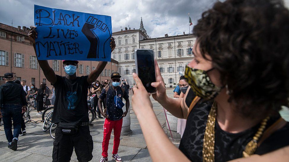 A woman wearing a protective mask photographs with a smartphone a man holding a Black Lives Matter poster