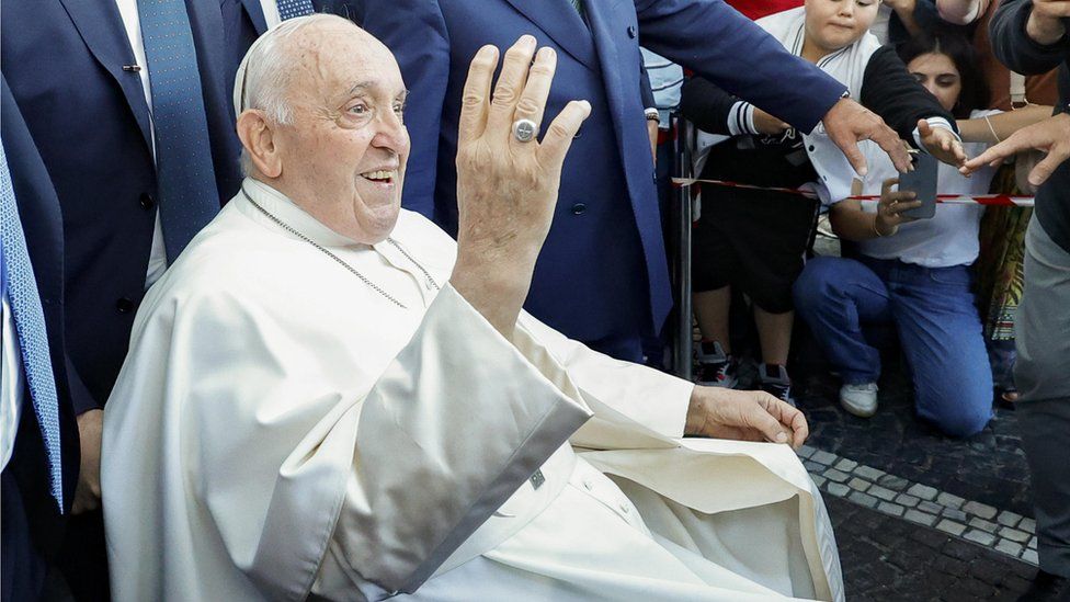 Pope Francis in a wheelchair smiling and waving to people as he leaves hospital in Rome