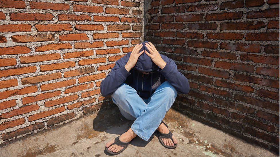 man sitting in corner with hoodie and hands over his head