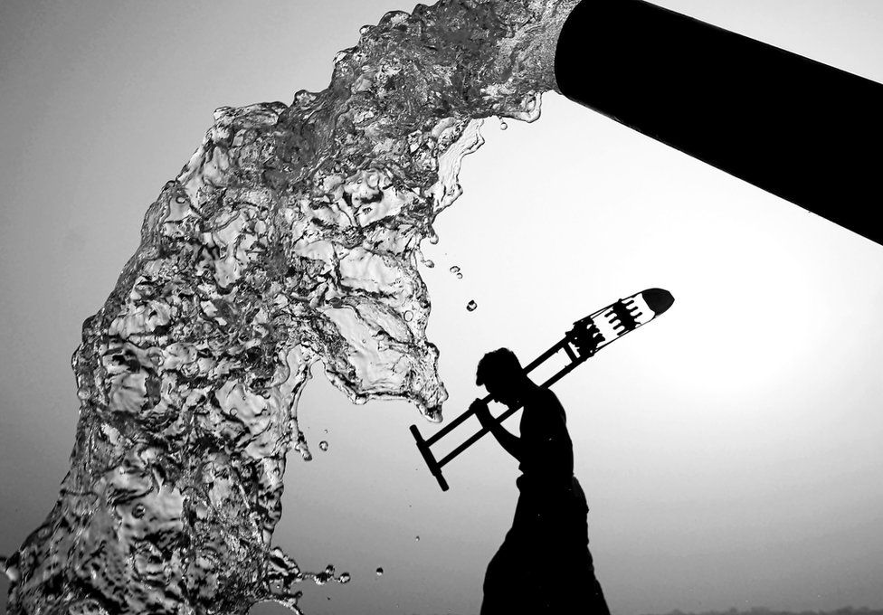 A black and white photo of a pipe gushing water with the silhouette of a man walking in the background