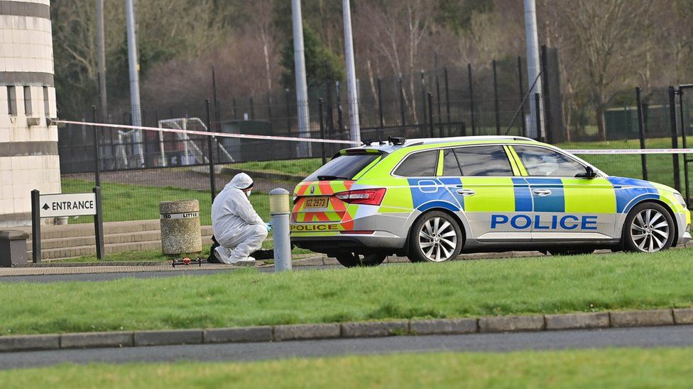 A forensics officer in a white suit crouches behind a police car parked at the scene of John Caldwell's shooting