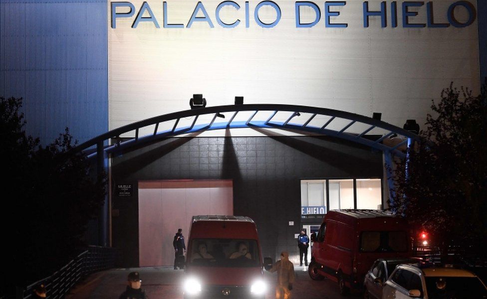 Police and military wearing protective suits stand as a van leaves the Palacio de Hielo shopping mall where an ice rink was turned into a temporary morgue on March 23, 2020