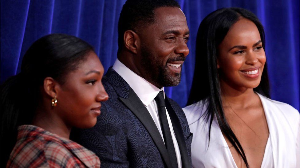 Idris Elba poses with his wife Sabrina and his daughter. All three are smiling against a blue backdrop. Idris is wearing a smart suit.