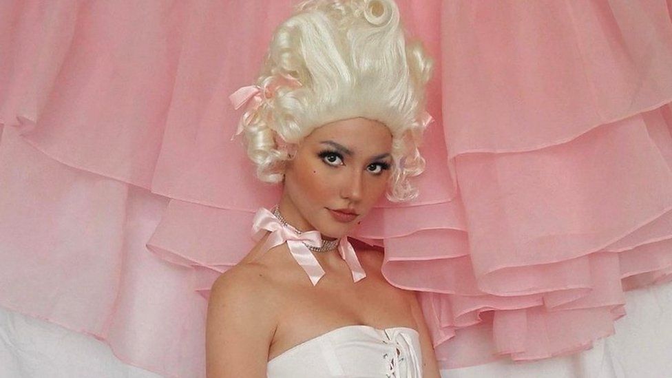 Influencer Janice Glimmer in a white curly wig styled to look like Marie Antionette wearing a corset and a pink bow and jewels around her neck