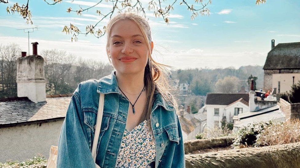 Jodie is an 18-year-old white woman, she wears her long blonde hair tied back with her pony tail pulled over her left shoulder. She wears a denim jacket over a floral dress with a canvas tote bag over her right shoulder and a charm necklace on a black string. She is smiling at the camera and is photographed outside, with the roofs of houses and chimneys visible behind her as well as a line of trees against a pale blue sky