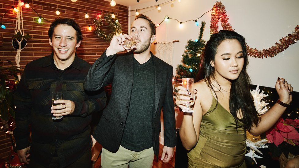 Generic image of three people at a Christmas party. There are two white men, dressed in smart-casual outfits, holding drinks and a woman in a green dress with long dark hair dancing. Behind them the room is decorated with fairy lights, tinsel and a Christmas tree