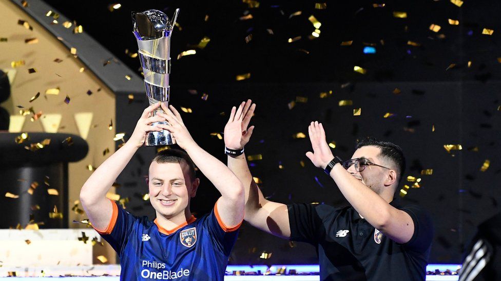 Manuel Bachoore celebrates winning the FIFAe World Cup 2023. He's holding the tall, cylindrical trophy on top of his head like a hat. He's smiling and wearing a navy blue football shirt with his team's logo on the left breast, and a New Balance logo on the right. His team captain Renzo is stood next to him, wearing the same t-shirt but in black, and he's holding his hands up as if bowing in worship. Gold pieces of confetti rain down on the stage around the pair