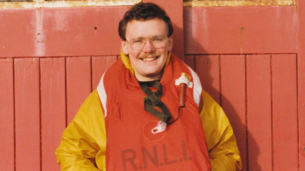 A man with a moustache and glasses wearing a yellow jacket and red life vest smiles at the camera