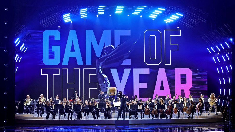 An orchestra performs on a large stage with "Game of the Year" projected on to a screen at the rear in colourful block capital letters. An image of the awards statue - a winged figure leaning backwards as if about to take flight - is superimposed on top. There are between 30 and 40 members of the orchestra, most of them seated and playing a range of stringed and woodwind instruments. To the far right, an electric guitarist stands facing the crowd, and a large cylindrical Japanese Taiko drum is on a plinth in the centre of the group.