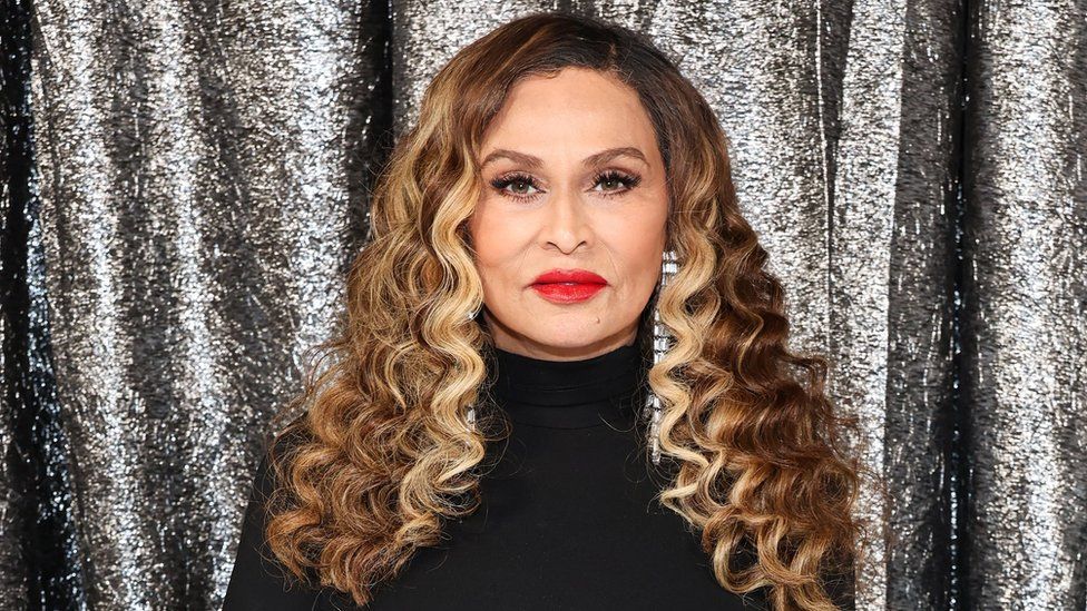 Tina Knowles attends the World Premiere of "Renaissance: A Film By Beyoncé" at Samuel Goldwyn Theater on November 25. Tina is a 69-year-old woman with brown hair and blonde highlights styled in ringlets just past her shoulders. She has brown eyes and wears a statement red lipstick. She's dressed in a high-necked black dress with long crystal earrings. She's photographed in front of a metallic silver backdrop.