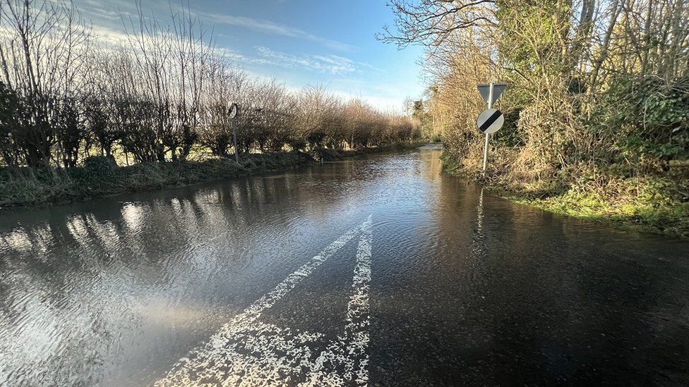 A flooded road in the Wiltshire countryside, hedges down the side with light reflecting off the water and a blue sky