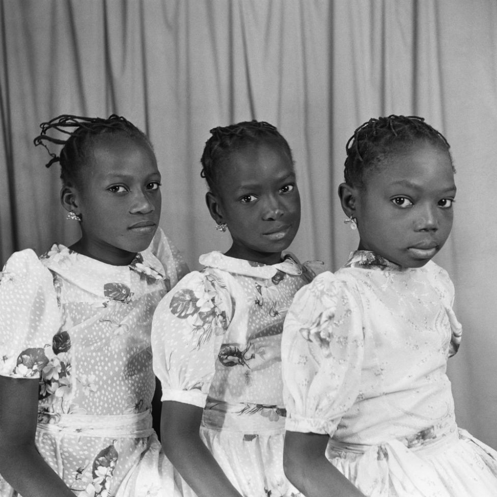 Three young girls wearing matching dresses, earrings and threaded hairstyles sit in a line and pose for the camera.