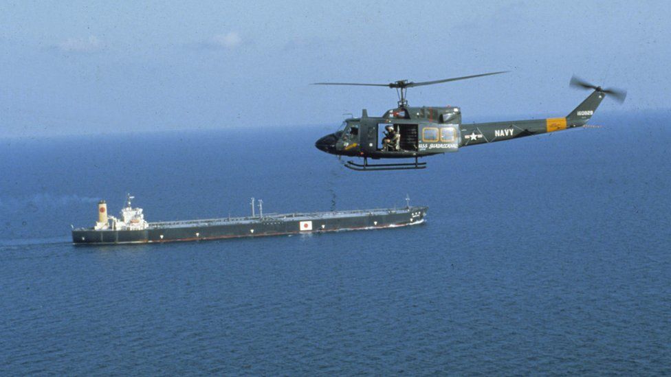 A green helicopter with its door open and soldiers visible flies above a tanker bearing what appears to be a Japanese flag - the helicopter bear the word NAVY on its tail and the roundel insignia of the US air force
