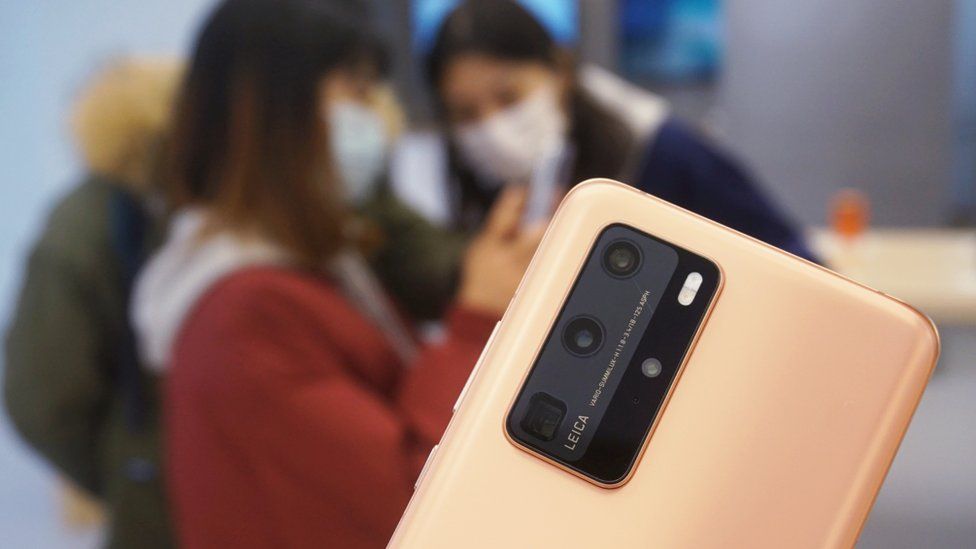 The rear panel of a Huawei P40 phone is seen in sharp focus, against a blurred background scene of customers trying out the phones