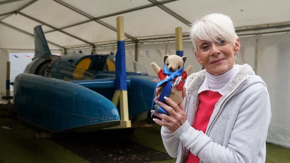 Gina Campbell standing in front of Bluebird at Bute in 2018, holding her father's mascot Mr Whoppit