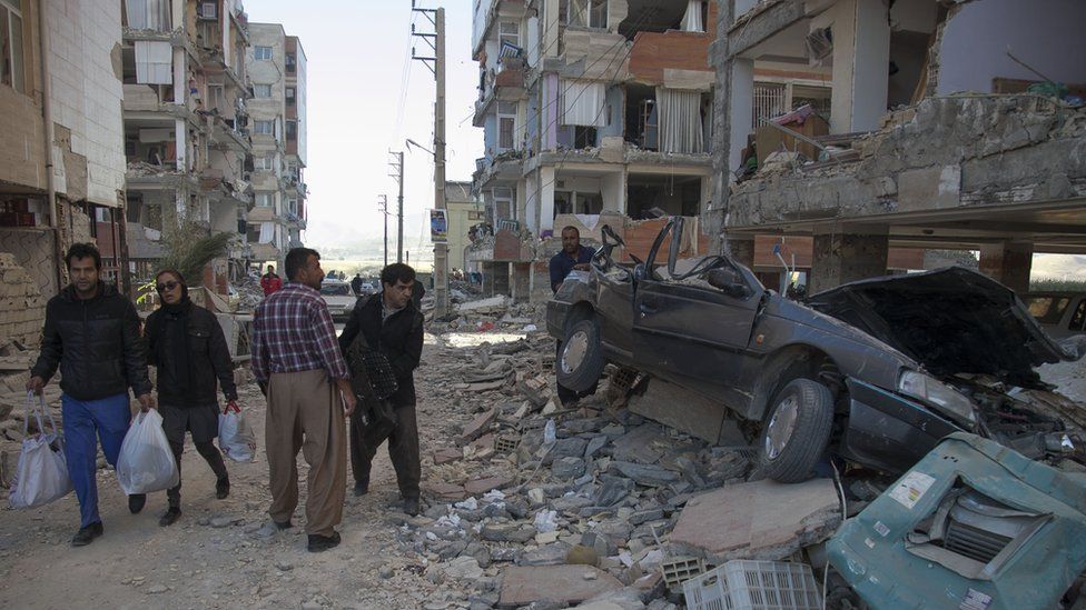 people walking past a car squashed under rubble in a street where parts of buildings have collapsed