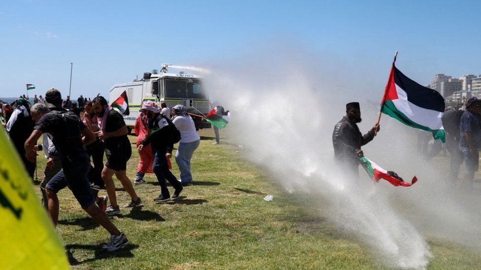 A crowd of protestors holding the Palestine flag are sprayed with a water canon in a field - Sunday 12 November 2023.