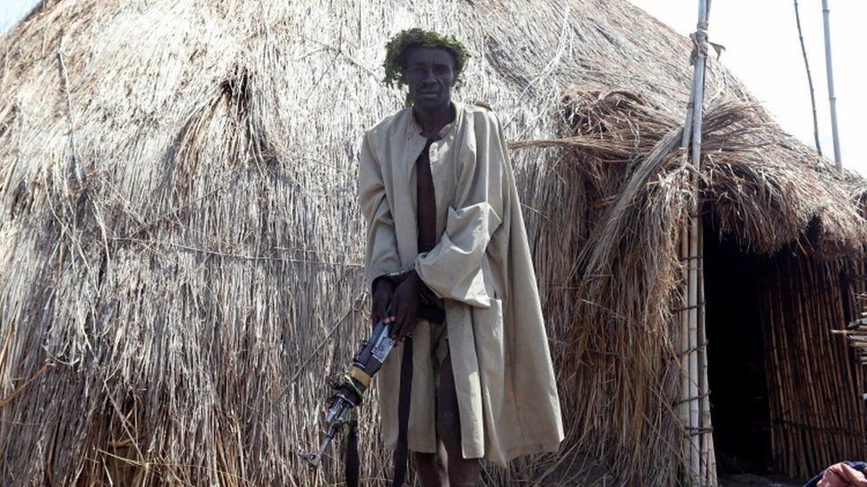 A man holding a rifle stands in front of a hut.
