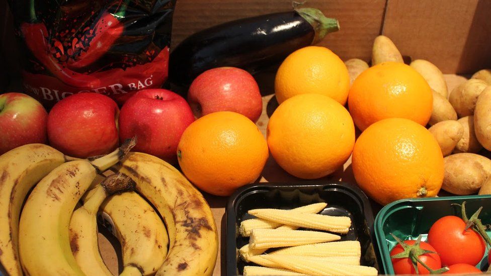 A box of fruit, vegetables and a packet of crisps