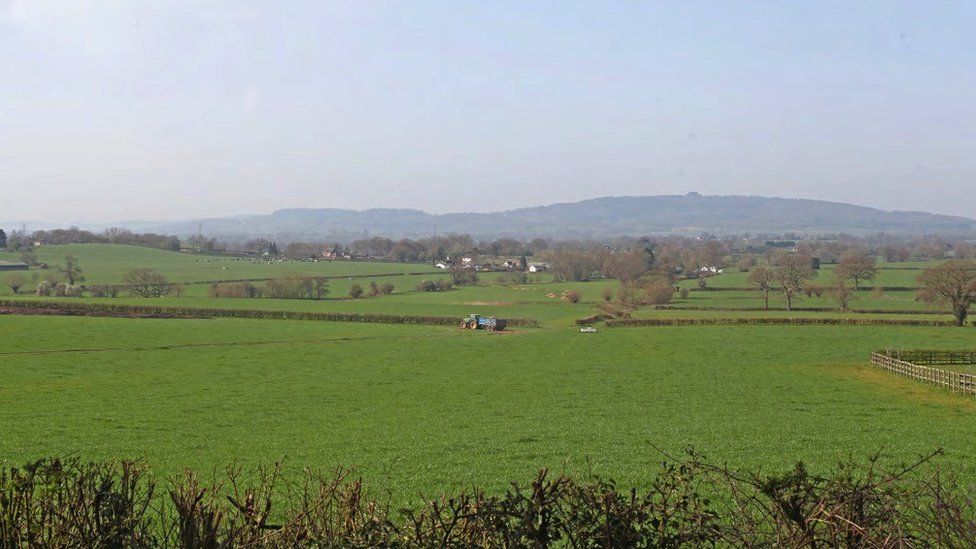 Image showing the land near Highleadon where JBM Solar Projects 21 Ltd wants to create a solar farm. There are very few houses on the large field and a blue tractor in the middle of it. More fields can be seen in the distance.