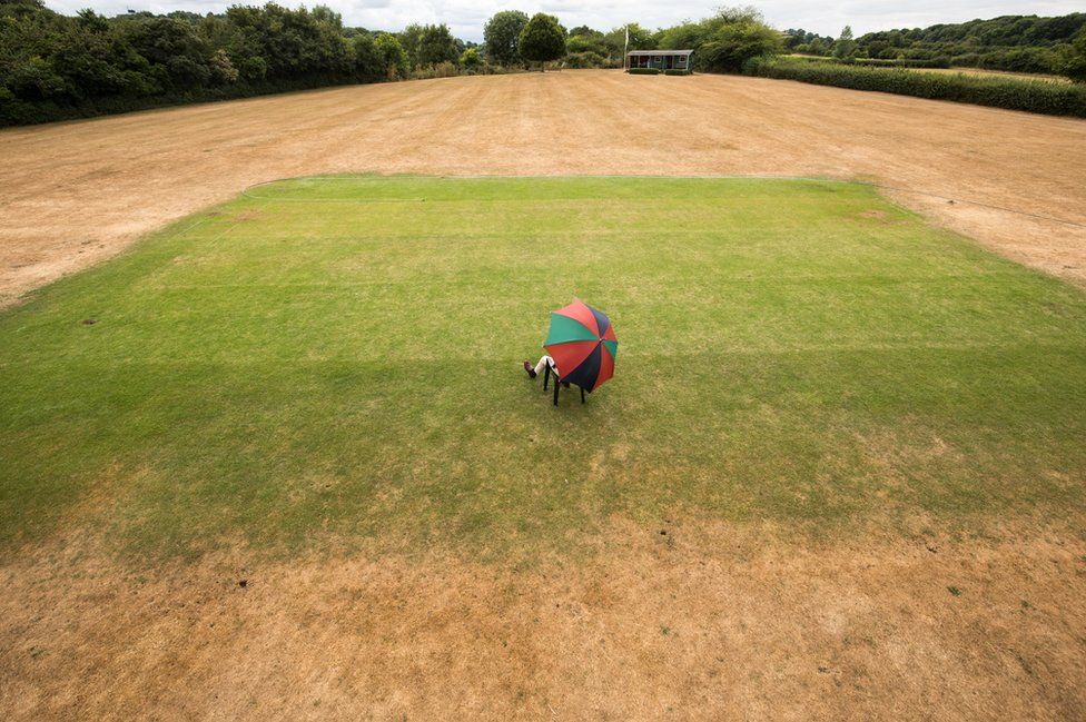 A man poses for a photograph at Priston Cricket Club as he sits on the wicket that has been watered by a hosepipe.