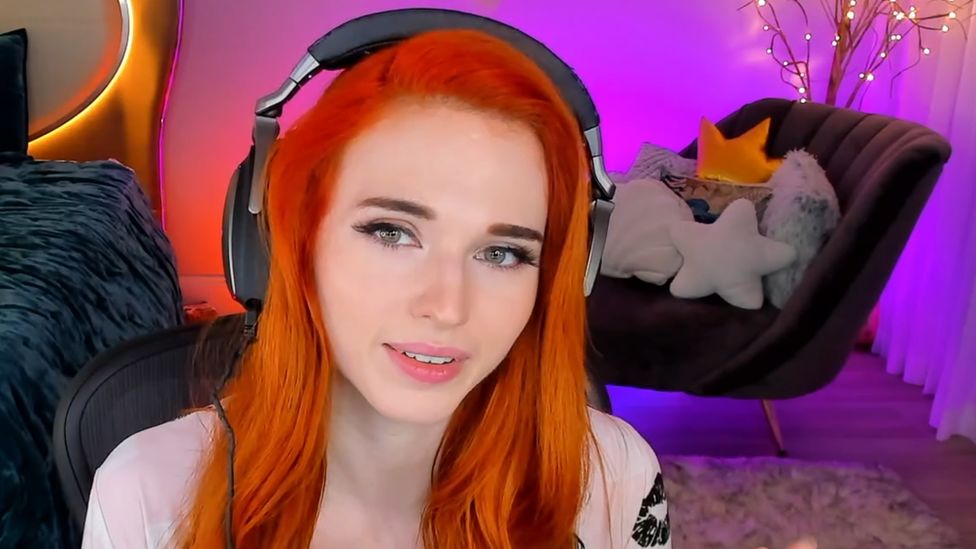 Amouranth, a young woman with red hair, wears headphones and looks at the camera in this screenshot from one of her streams