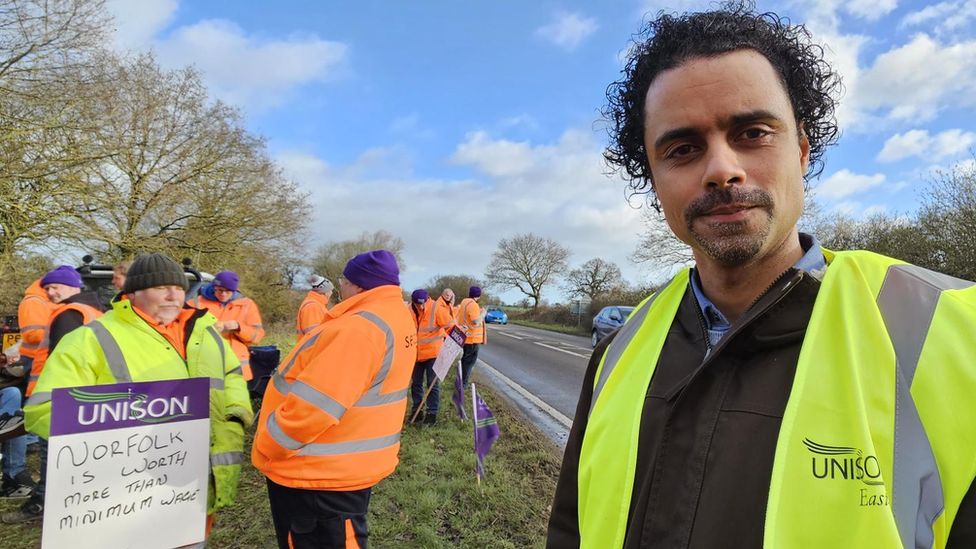 Cameron Matthews wearing a hi-vis yellow waistcoat standing beside the A140 a man next to him holds a sign which says Unison: Norfolk is worth more than minimum wage