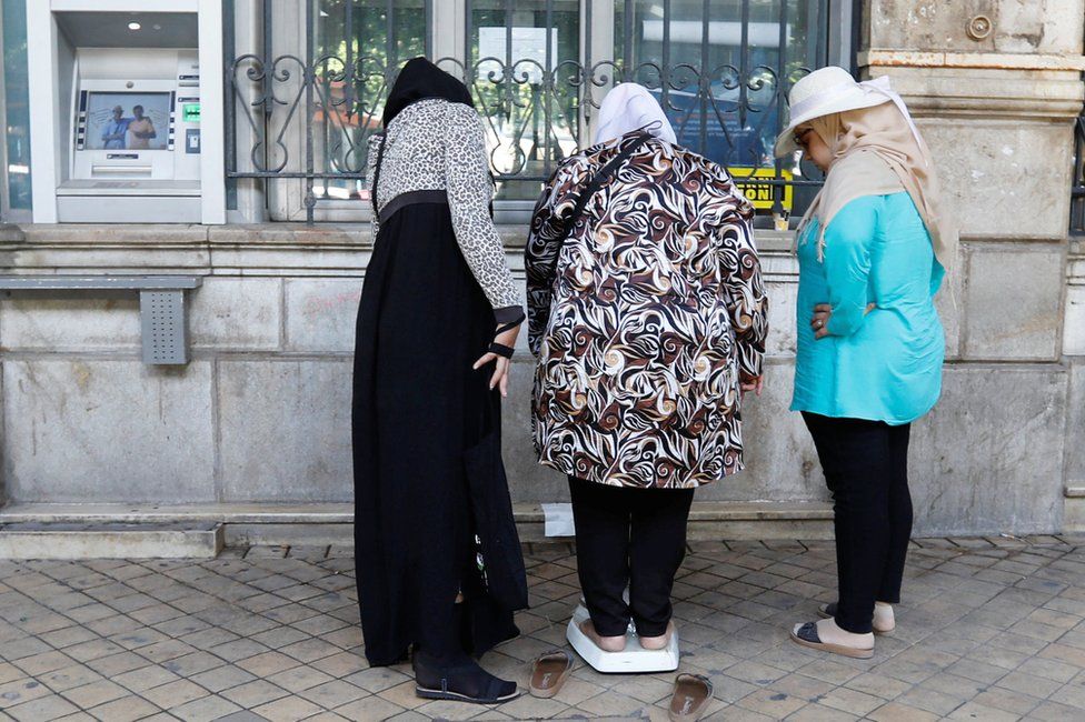 A woman on a weighing scale outside a bank observed by two other women in Tunis, Tunisia - Thursday 30 August 2018